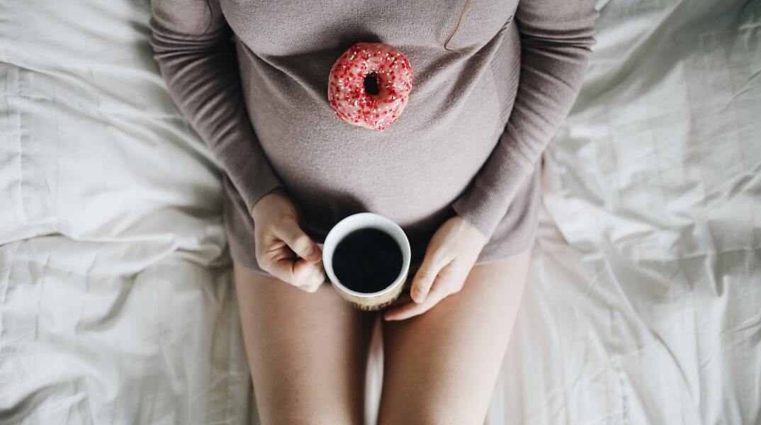 Εγκυμονούσα ξαπλωμένη στο κρεβάτι πίνει ρόφημα που μοιάζει σε καφέ και έχει στηρίξει στην κοιλίτσα της ντόνατ