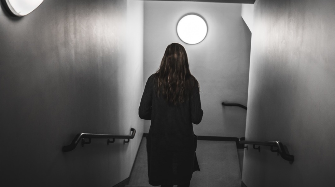 Γυναίκα περπατά με την πλάτη γυρισμένη στον φακό προς πόρτα που βρίσκεται σε κατηφορικό σκοτεινό διάδρομο