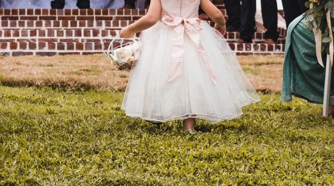 Κοριτσάκι με φόρεμα που παραπέμπει σε γάμο διασχίζει σημείο με γρασίδι και κρατά στα χέρια του καλαθάκι