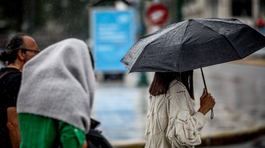 Γυναίκα με ομπρέλα προστατεύεται από τη βροχή - Άτομο έχει ρίξει ρούχο στο κεφάλι του για να μην βραχεί