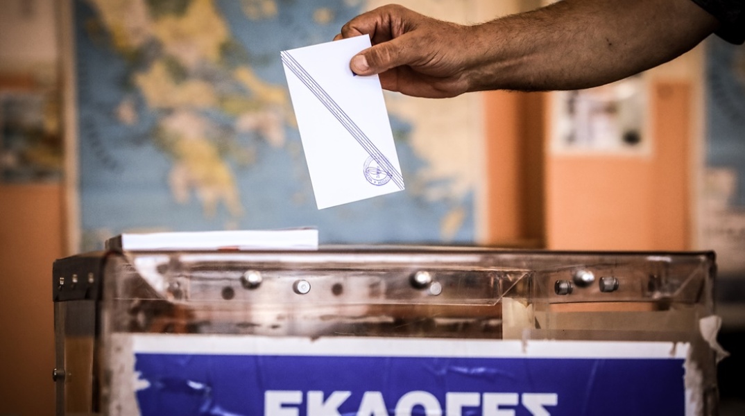 Χέρι με ψηφοδέλτιο πάνω από την κάλπη και φόντο τον χάρτη της Ελλάδας κρεμασμένο σε τοίχο σχολικής τάξης