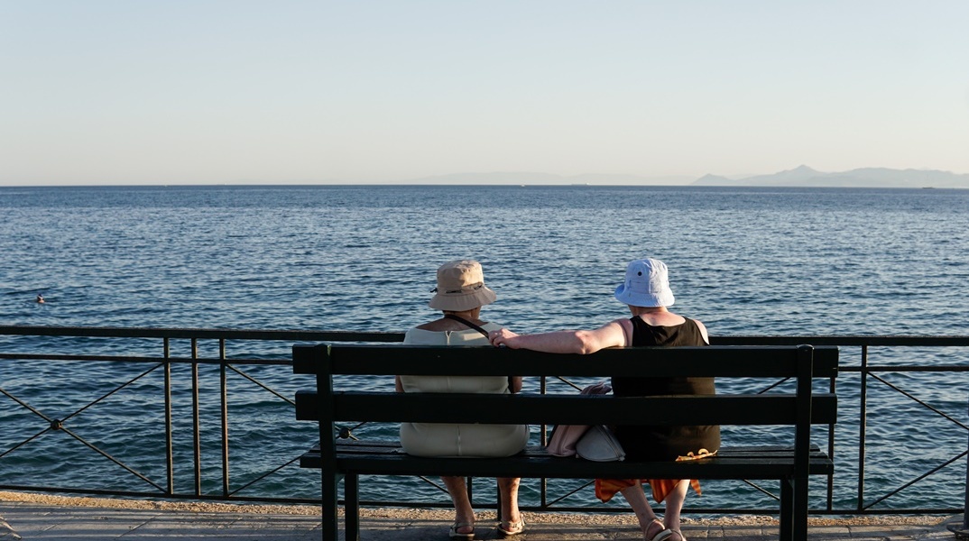 Ηλικιωμένες κυρίες κάθονται φορώντας τα καπελάκια τους σε παγκάκι με θέα τη θάλασσα