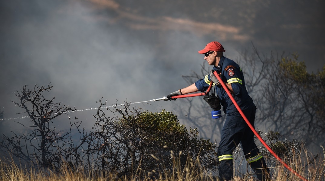 Πυροσβέστης με μάνικα ρίχνει νερό σε περιοχή όπου έχει περάσει φωτιά για τον κίνδυνο αναζωπυρώσεων