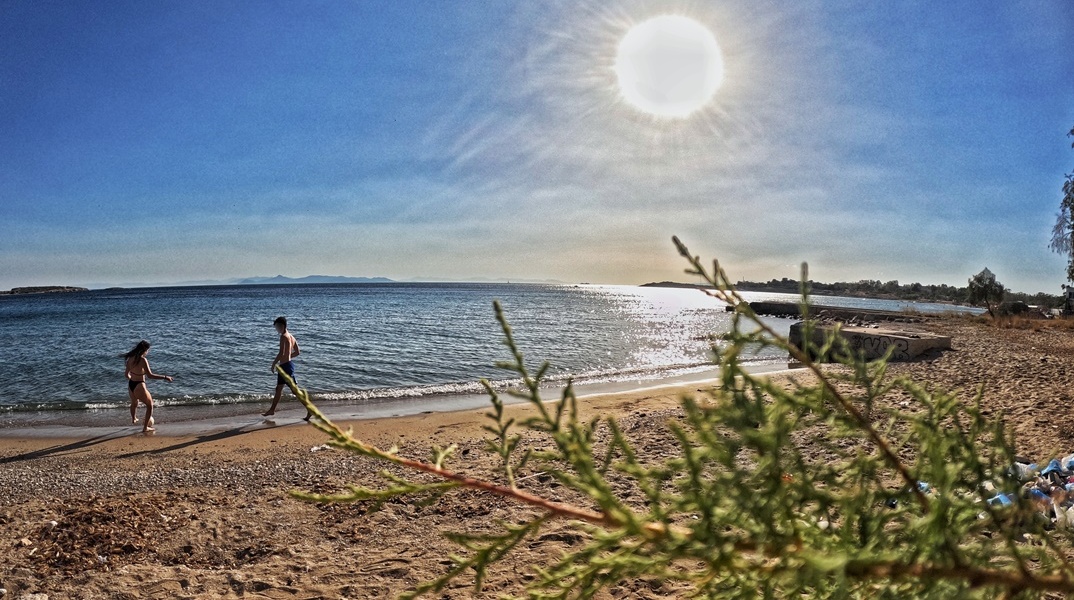 Ακτίνες του ήλιου πέφτουν στον φωτογραφικό φακό ενώ τραβά σκηνή από παραλία με αμμουδιά και δύο άτομα με μαγιό να βρίσκονται στην ακρογυαλιά