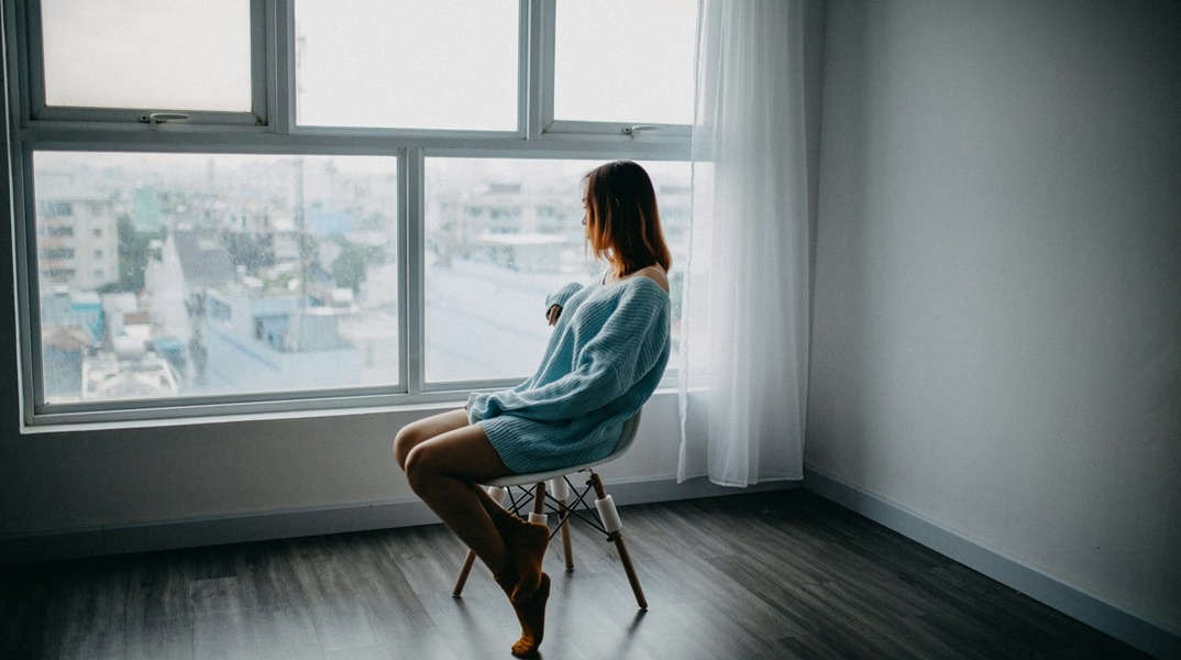 Γυναίκα λυπημένη κάθεται σε καρέκλα άδειου δωματίου και κοιτά από το παράθυρο