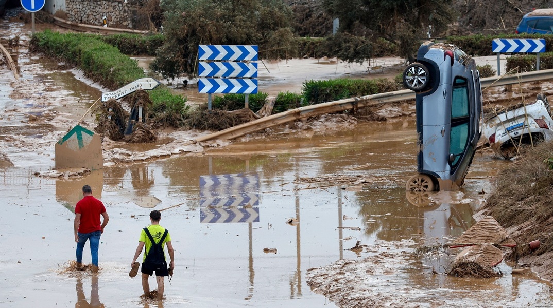 Εικόνα από δρόμο της Βαλένθια μετά τις φονικές πλημμύρες στην Ισπανία: Πολίτες περπατούν στη λάσπη και αυτοκίνητο που έχει παρασυρθεί στέκεται όρθιο